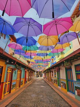 Colorful umbrellas hanging over narrow cobblestone pathway, illuminating scenic street corner in guatape, colombian landmark, full of colorful houses decorated with bas-relief designs (zocalos)