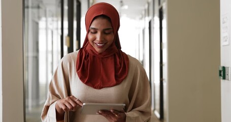 Focused Arabian businesswoman dressed in Muslim hijab and abaya standing in modern office corridor using digital tablet. Technology usage in workflow, productivity, women in business across cultures - Powered by Adobe