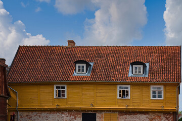Historic building with a yellow wooden facade and red tiled roof under a bright blue sky. Dormer windows with white frames and brick chimney add charm to the traditional European architecture.