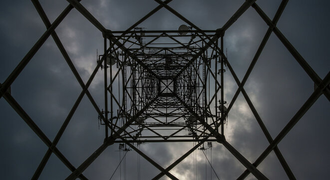 A low angle shot of a metal transmission tower against a cloudy sky showing geometric patterns upward