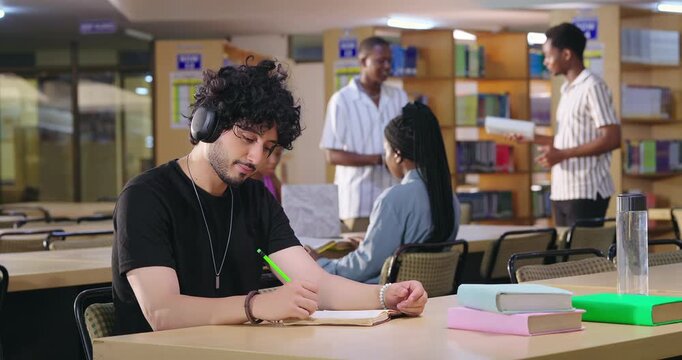 Indian asian young adult gen z male man calm peace mind student sitting desk table listening music hold pencil write exam notes smart boy do self study read book prepare topics indoor library busy day