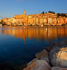 Panoramic sunset view of Menton, France, with colorful old town buildings glowing in warm light, set against the Mediterranean Sea and blue sky on the French Riviera