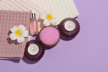 Folded waffle towels, burning candles, spa products and plumeria flowers on violet background, flat...