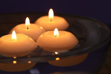 Burning candles in bowl of water on dark purple background, closeup