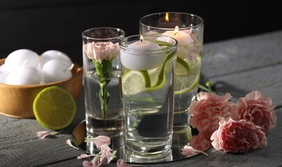 Glasses of water with candles, flowers and lime on grey wooden table, closeup
