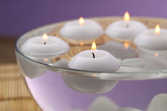 Burning candles in bowl of water on table against purple background, closeup