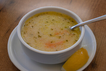 Close-up of steaming Turkish soup in a white bowl with visible herbs and carrot slices, accompanied by a lemon wedge, on a wooden table. Perfect for illustrating traditional cuisine and comfort food.