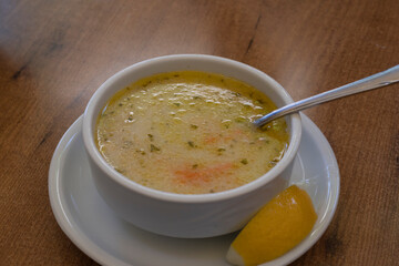 Close-up of steaming Turkish soup in a white bowl with visible herbs and carrot slices, accompanied by a lemon wedge, on a wooden table. Perfect for illustrating traditional cuisine and comfort food.