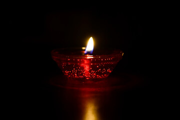 A red oil lamp or Diya with a lit flame, isolated against a completely dark background.