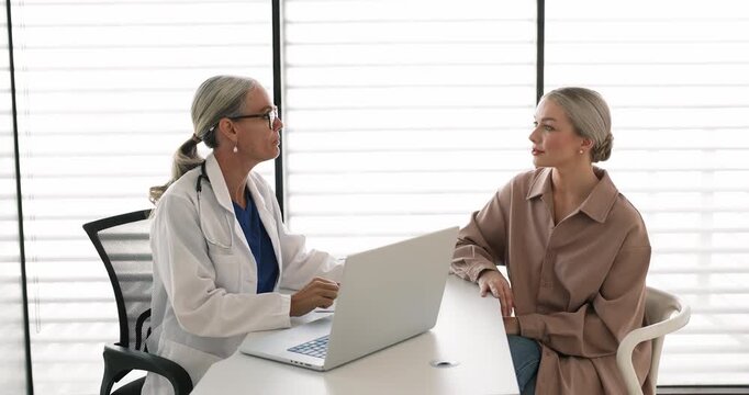 Young attractive woman patient shares health-related concerns, ask questions, explain symptoms, speaks to experienced mature female doctor during visit at hospital. Professional healthcare services