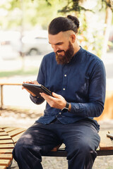 A man with a groomed beard sits on a park bench, focused on his tablet in a peaceful outdoor setting