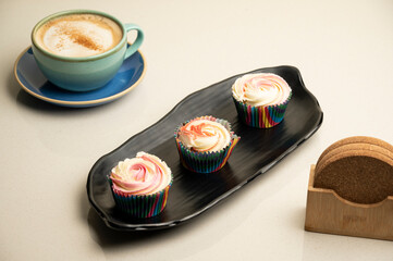 Aesthetic shot of strawberry, vanilla, and pineapple cupcakes on a black plate, served with cappuccino coffee in a mint green cup on a blue plate with wooden coasters, all set against white background
