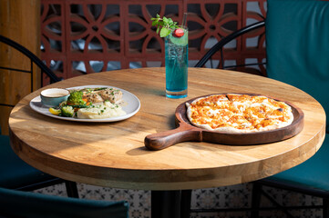 Chicken steak with creamy pepper sauce, sautéed vegetables, mashed potatoes, and hot sea pizza topped with prawns, calamari, and fish, served with blue cocktail on wooden table in sunlight.