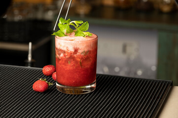 Close-up shot of strawberry-flavoured drink in a highball glass on bar mat, with fresh strawberries nearby and bartender placing mint garnish with tongs, blurred bar background