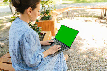 A young woman uses her laptop in nature, blending productivity with tranquility in a serene...