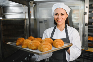Baker woman holding tray of freshly baked croissants in bakery kitchen