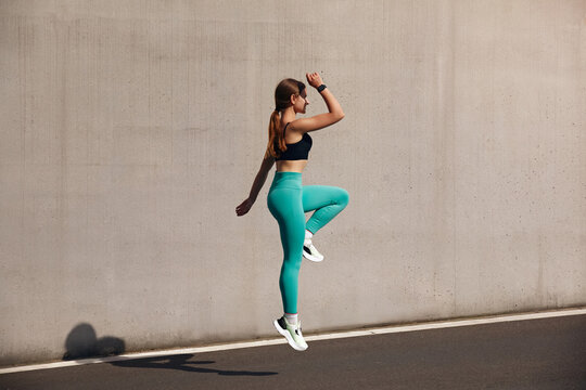 Fit woman training outdoors, in an urban environment. Healthy young sportswoman doing fitness. An athlete makes jumps, does functional training on the street on a bright sunny day, smiles contentedly - Powered by Adobe