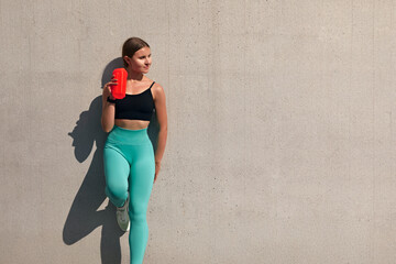 Young athlete with hand on hip drinking water in front of wall. A girl quenches her thirst on a sunny day. Maintaining hydration
