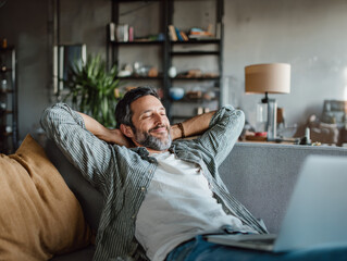 Fototapeta premium Relaxed freelancer leaning back on a couch with a laptop in front of him—perfect for illustrating remote work freedom, work-life balance, or home office comfort.