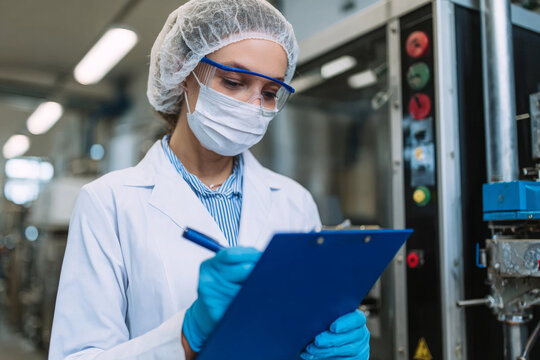 Female factory worker in protective gear writing on clipboard near machinery.