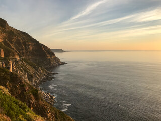 Rocky coastal cliffs at golden sunset, Cape Town. A serene ocean view with rugged rocky cliffs, calm waves, and a warm golden sunset casting light across the tranquil seascape.

