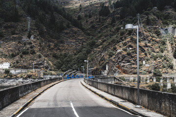 Roadway across a concrete dam with blue floodgates, flanked by steep rocky hillsides with power...