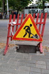 A Worn Triangular "Road Work" Warning Sign On A Wooden Barrier In Front Of An Open Manhole On A City Sidewalk. Urban Danger And Repair Concept. Vertical.