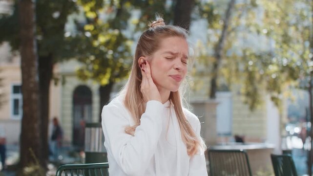 Earache. Sick Caucasian young woman suffers from ear pain sitting on chair in city street. Upset girl teenager touches her ear. She has furuncle, shingles, eczema, ear diseases, or otitis media.