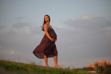 Graceful pregnant woman posing on grassy hilltop with flowing dress under colorful sunset evening...