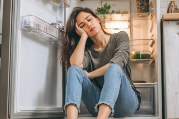 Exhausted Woman Sitting on Kitchen Floor by Empty Refrigerator