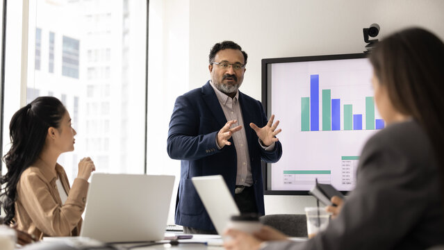Corporate training. Experienced mid aged business coach trainer lead education event at modern office boardroom for multicultural young workers team explain data analysis to trainees using infographic