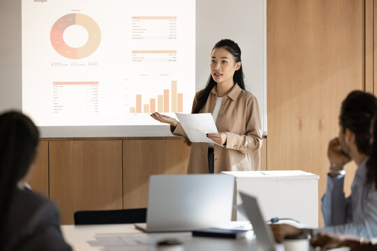 Fototapeta Data driven seminar. Young Asian woman speaker lead interactive presentation in modern meeting room use digital screen to visualize project information in charts graphs give report to diverse audience