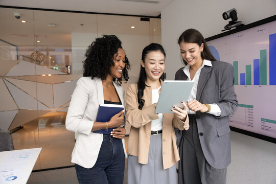 Happy smiling young project manager social media marketer share idea with multicultural female colleagues teach teammates to work in corporate app on pad show good results of collaborative research