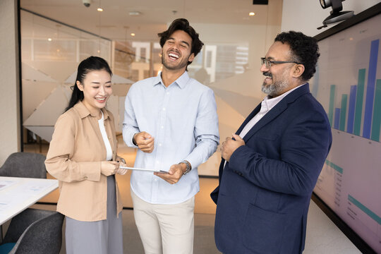 Business fun. Overjoyed international team of three diverse employees executives stand in office workplace with digital pad laughing at funny mistake share happiness of successful project achievement - Powered by Adobe