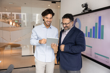 Product or service demonstration. Confident millennial manager project developer stand in modern office explain new business offer solution to interested mature male client colleague using tablet pc
