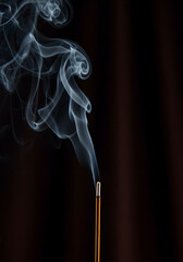A close up of incense stick burning with smoke curling against a dark brown background studio shot