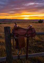 A saddle resting on a wooden fence with a sunset in the background over a grassy landscape