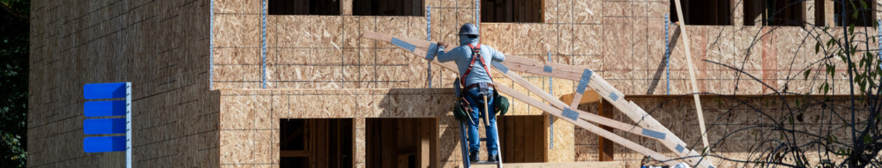 Carpenter on ladder with hard hat, tool belt, and safety harness pulling up a wood frame rafter...