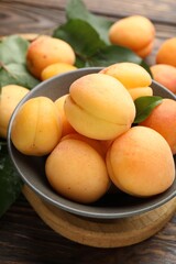 Fresh apricots in bowl and green leaves on wooden table, closeup