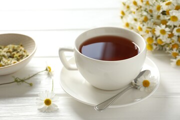 Aromatic tea in cup, chamomile flowers and spoon on white wooden table, closeup