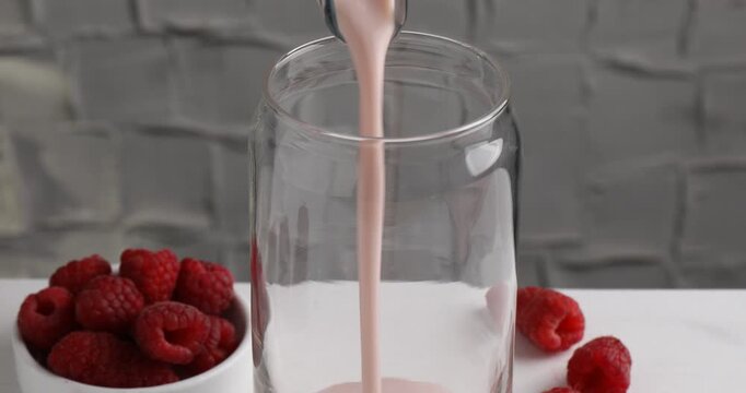 Pouring tasty raspberry milkshake into glass on white table, closeup