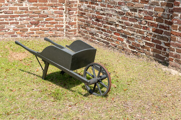 Wooden Wheelbarrow by Brick Wall. Vintage wooden wheelbarrow with metal wheel resting on grass...