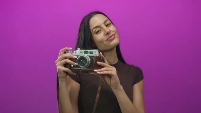 Woman posing with vintage camera in front of vibrant pink background showcasing a playful expression with long hair and casual outfit.