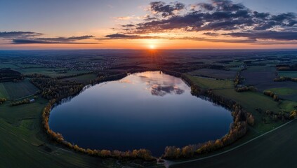 Aerial view of a serene lake at sunset.  A tranquil, circular lake, nestled within a landscape of farmland and trees, is bathed in the warm hues of a setting sun.  