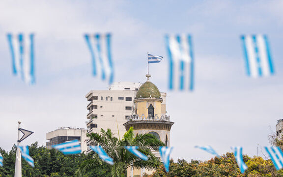 Mudejar-style clock tower with Honduran flag and festive decorations. Urban view with palm trees and blurred people in foreground. Guayaquil - Powered by Adobe
