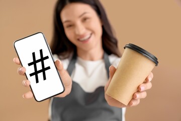 Female barista holding cup of coffee and mobile phone with hashtag sign on screen against beige background, closeup