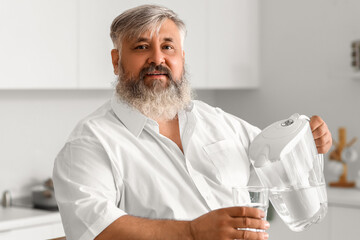 Mature man pouring purified water into glass from filter jug in kitchen