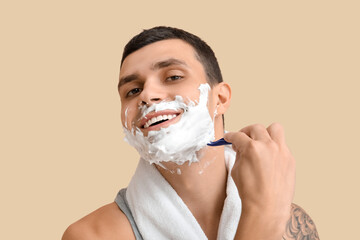 Handsome young man with towel and razor shaving his face against beige background