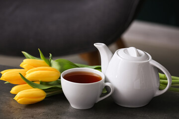 Cup of hot brewed tea, pot and tulips on table against black blurred background
