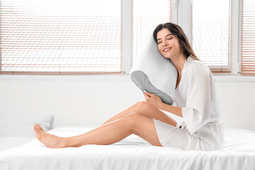 Young woman in pajamas holding soft orthopedic pillow on bed in room